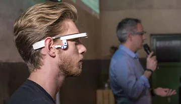 Joven con gafas inteligentes escuchando a un ponente con micrófono en una demostración tecnológica